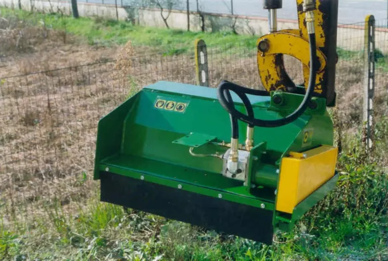 La trincia montata sul braccio di una macchina operatrice gialla, con i tubi idraulici collegati al motore, accanto a una recinzione.