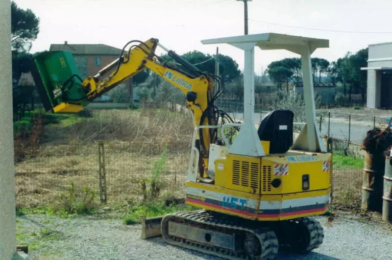 La trincia montata in cima al braccio di un miniescavatore giallo, fermo in un piazzale.