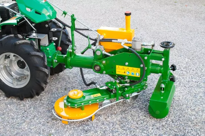 Erpice interfilare ERPI 1 verde con serbatoio idraulico giallo, montato su un motocoltivatore verde su ghiaia: si vedono il braccio fresa con il disco giallo, l’asta tastatore e il rullo anteriore.