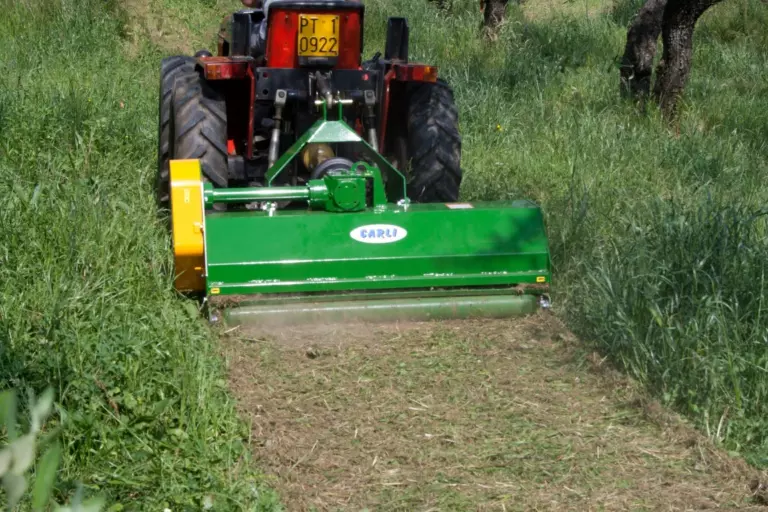 La trincia montata dietro un trattore rosso, al lavoro nell’erba alta tra gli alberi, con la striscia di erba trinciata dietro.