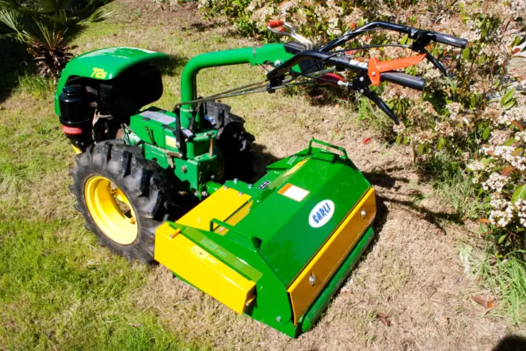 La trincia montata su un motocoltivatore verde, ferma sull’erba secca accanto a una siepe fiorita.