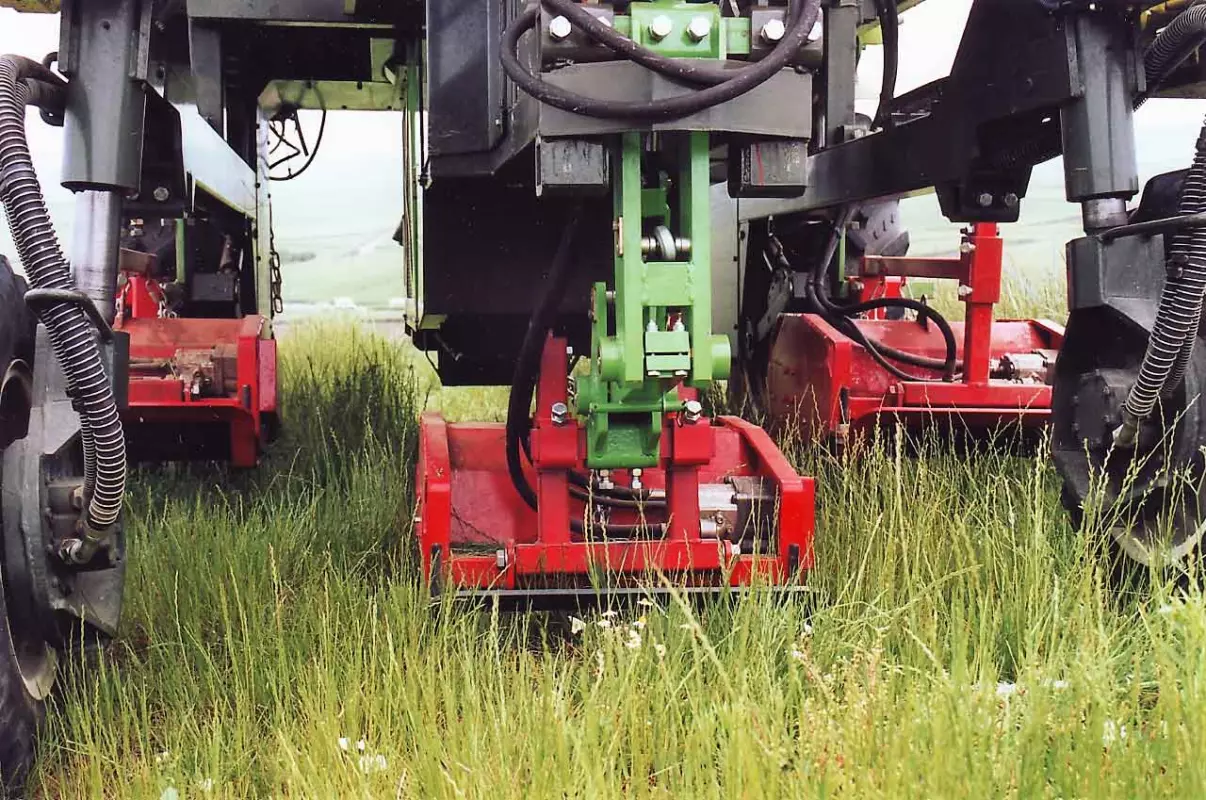 Three red flail mowers mounted under the frame of a vineyard machine, lowered into tall grass: the central one hangs from a green support.