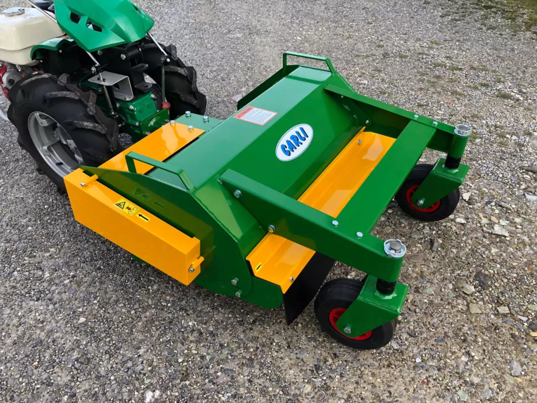 Trincia Professionale 118 verde e gialla con le ruote pivotanti a richiesta, montata su un motocoltivatore verde, su un piazzale di ghiaia.