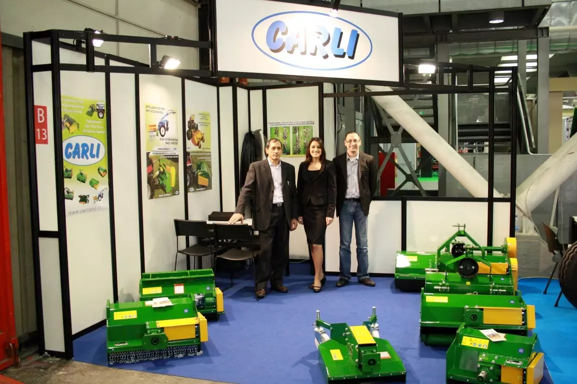 Three members of the Carli staff at the company’s trade fair stand, among green and yellow flail mowers